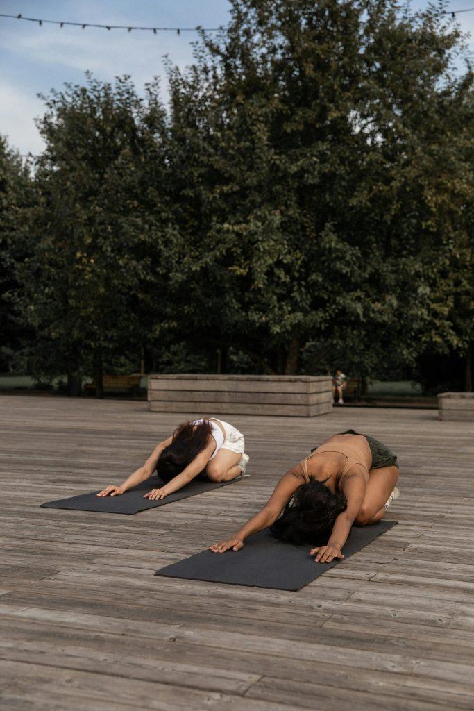Dos mujeres realizando yoga, en un suelo de madera, con un fondo con árboles.