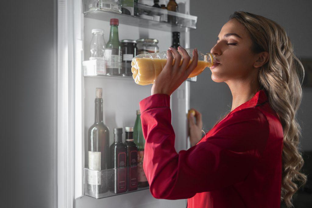 Una chica bebiendo zumo de naranja directamente de la botella de plástico sacada en la nevera.